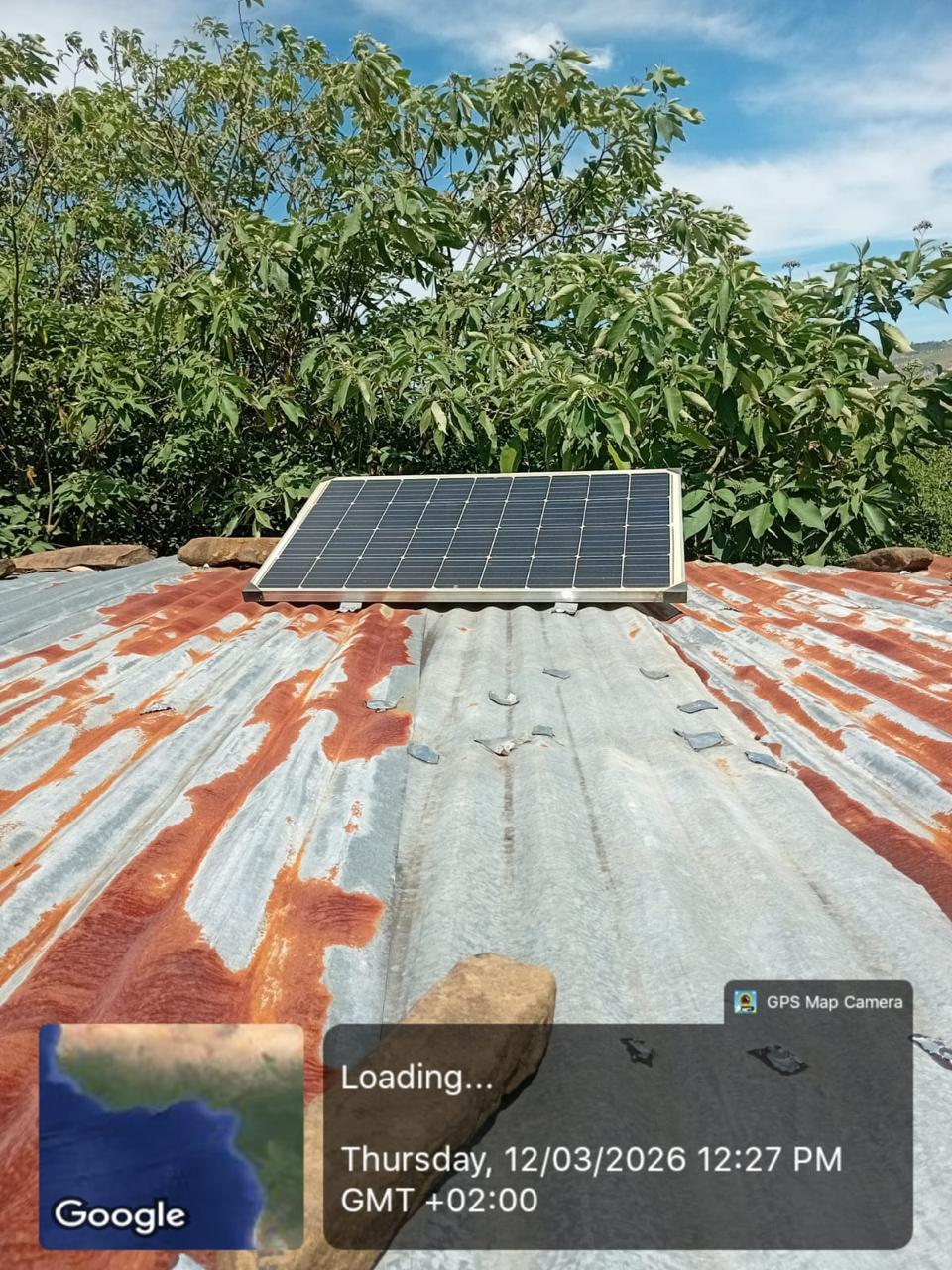 Solar panel mounted above a roof with nearby vegetation in the background.